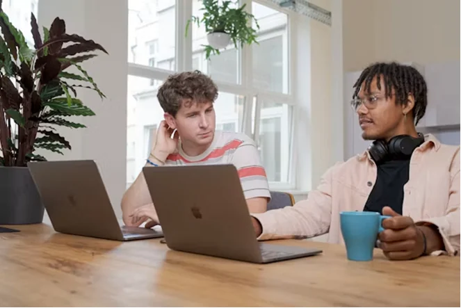 Deux jeunes adultes qui échangent à une table de travail, ordinateurs ouverts